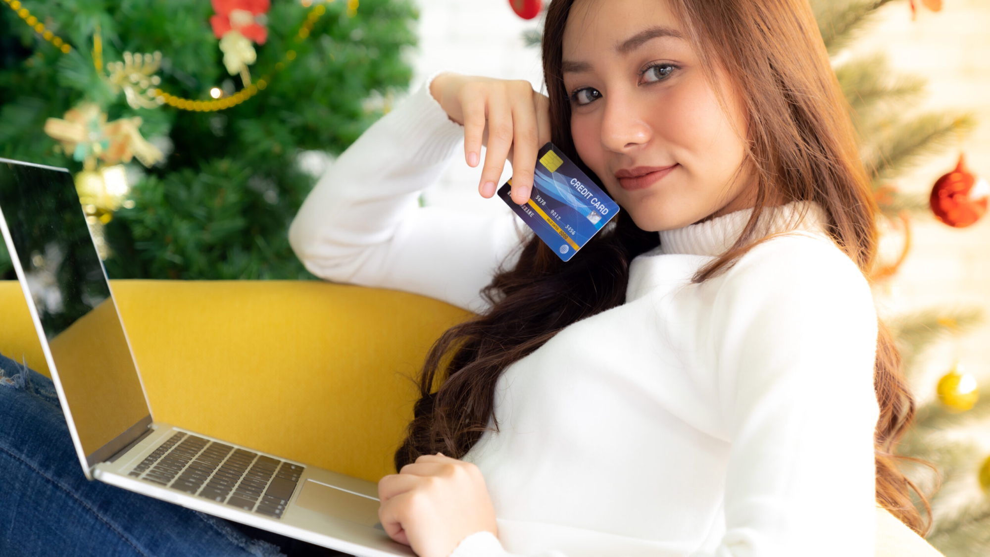 Young Adult teenager using laptop on sofa in living room for online shopping with gift box with Christmas tree in background