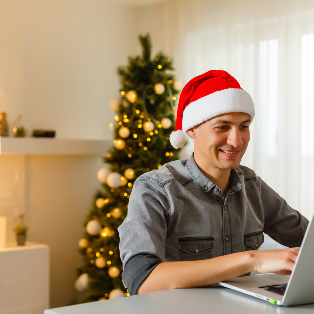 Man in Santa Claus hat, sitting in front of Christmas tree, searching on laptop.